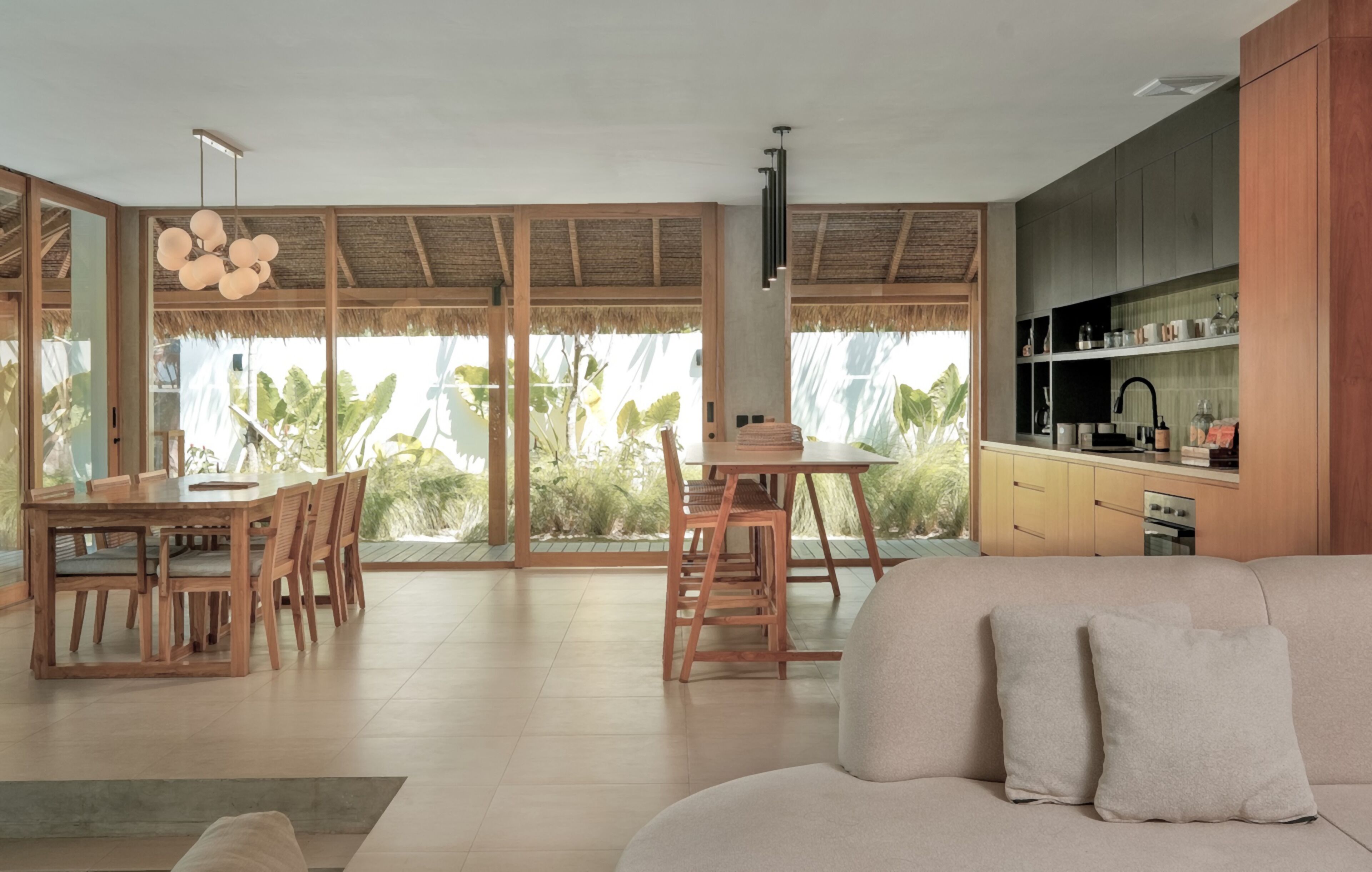 A spacious living area with a sofa, dining table, and chairs. There is a kitchen area with cabinets and a sink. Large windows provide views of greenery outside.