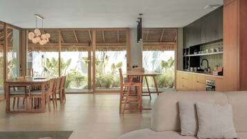 A spacious living area with a sofa, dining table, and chairs. There is a kitchen area with cabinets and a sink. Large windows provide views of greenery outside.