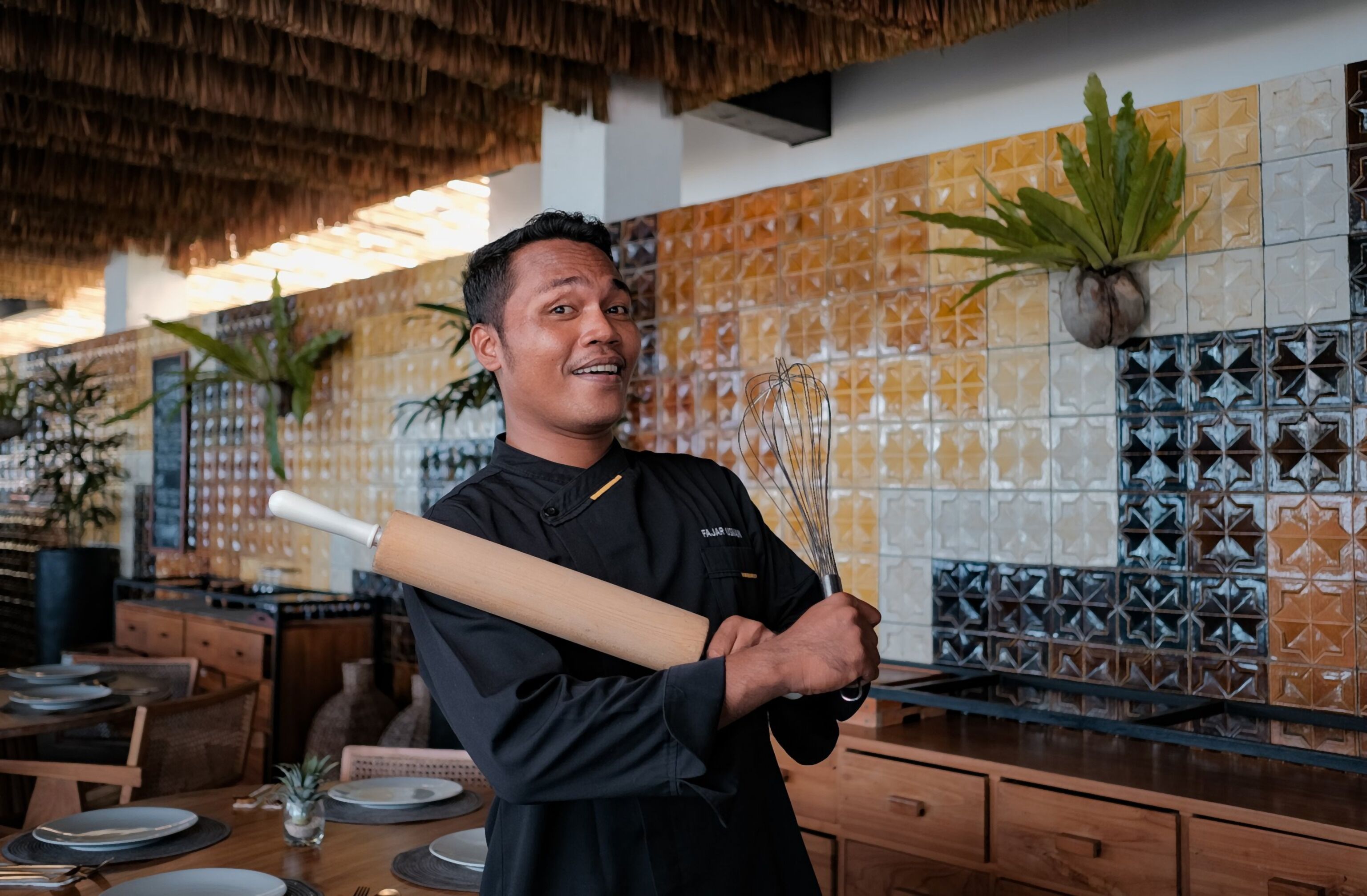 A chef in a kitchen with a thatched ceiling, wooden cabinets, and a tiled backsplash with geometric patterns.