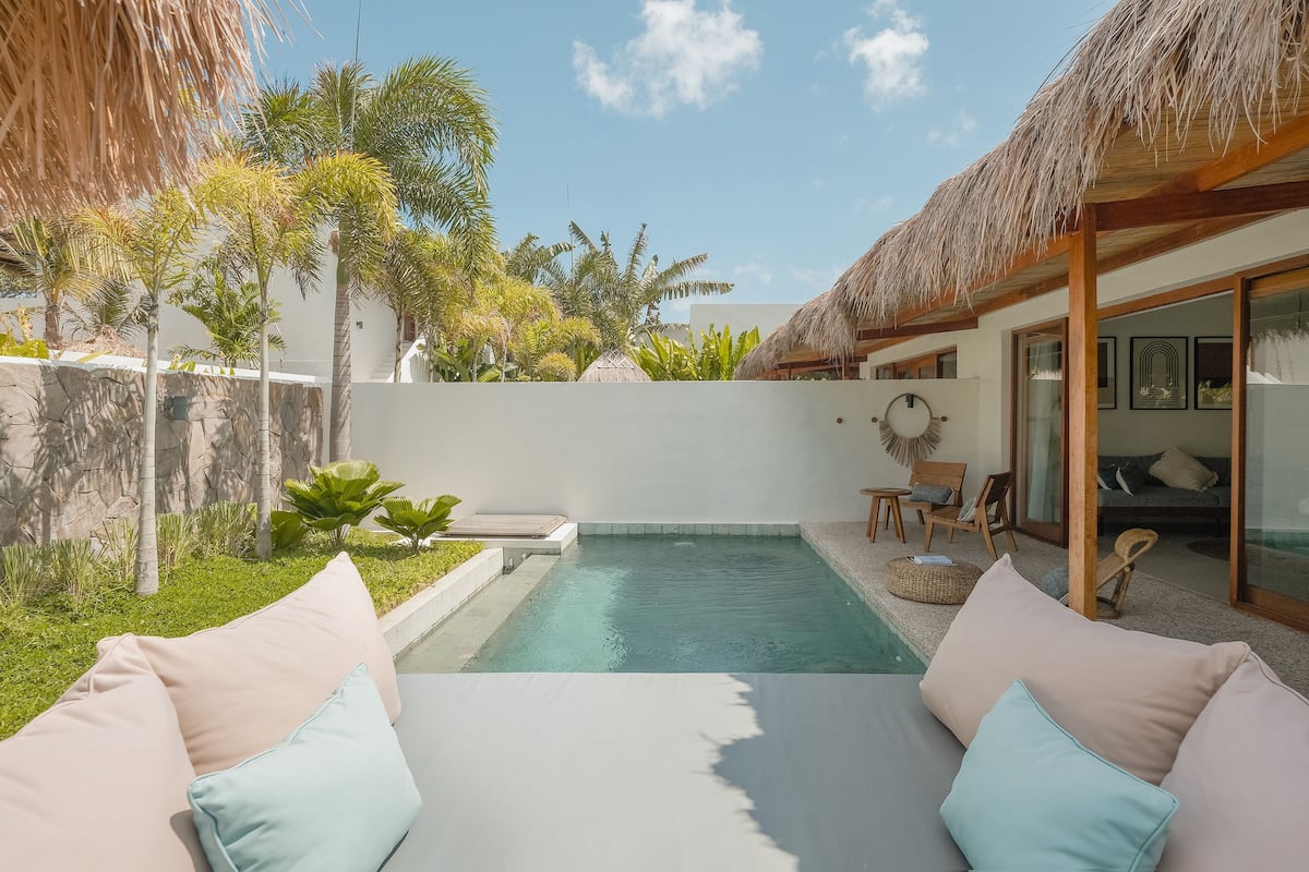A pool area with a thatched roof structure, a seating area with cushions, and a view of a modern building with glass doors.