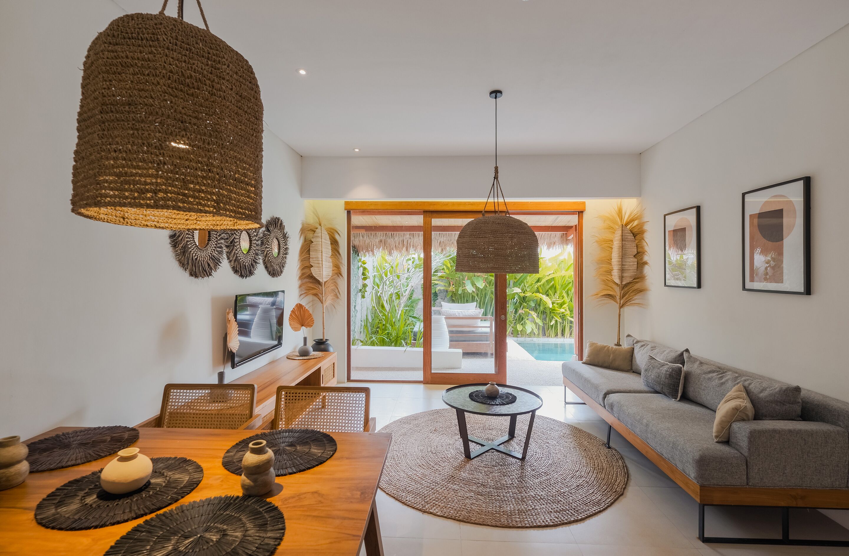 A modern living room with a wooden dining table, a grey sofa, and a large window overlooking a garden.