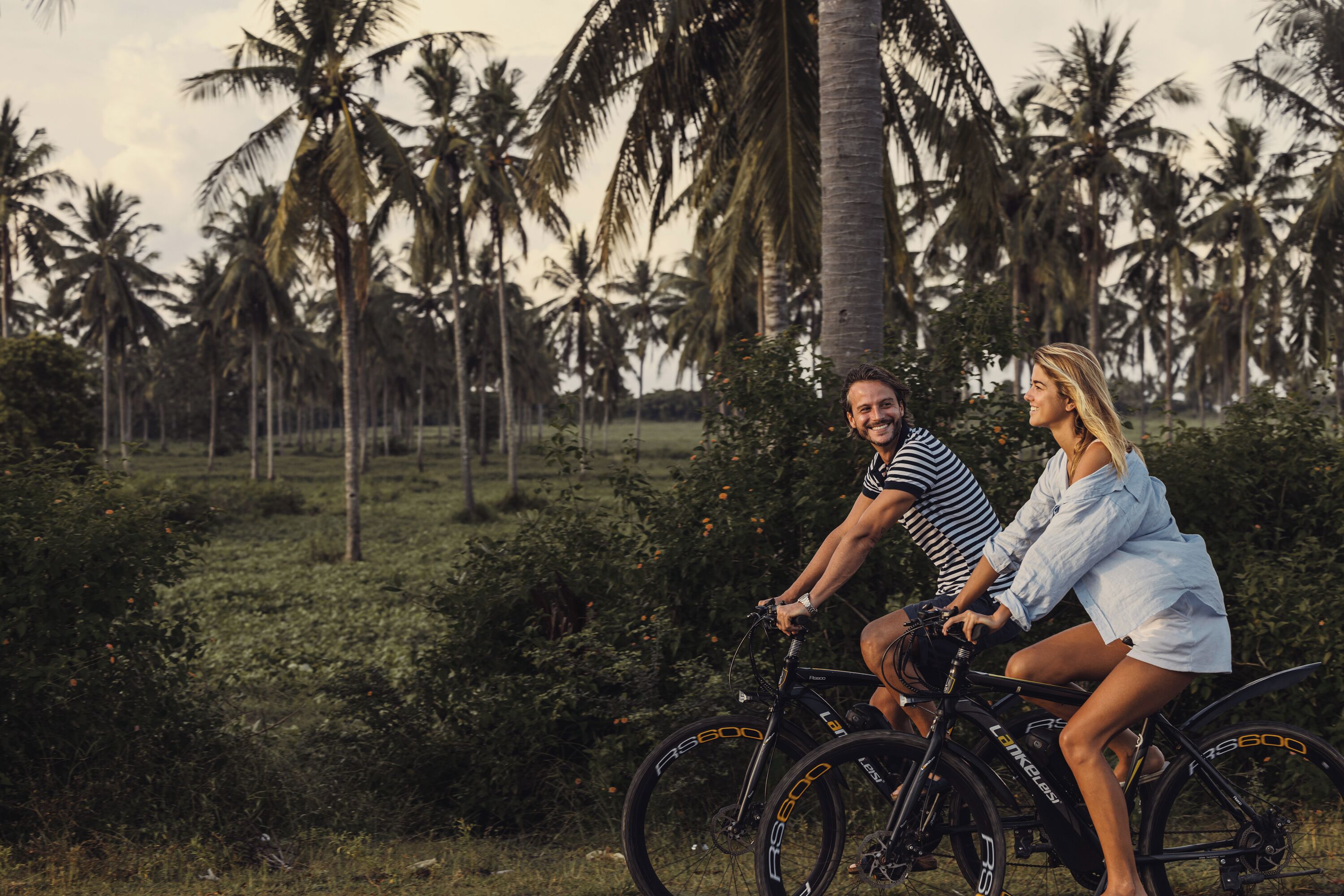 Two people on a bicycle in a tropical setting with palm trees.