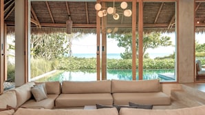 Una sala moderna con un sofá beige grande, una lámpara colgante y vistas a una piscina y al océano.