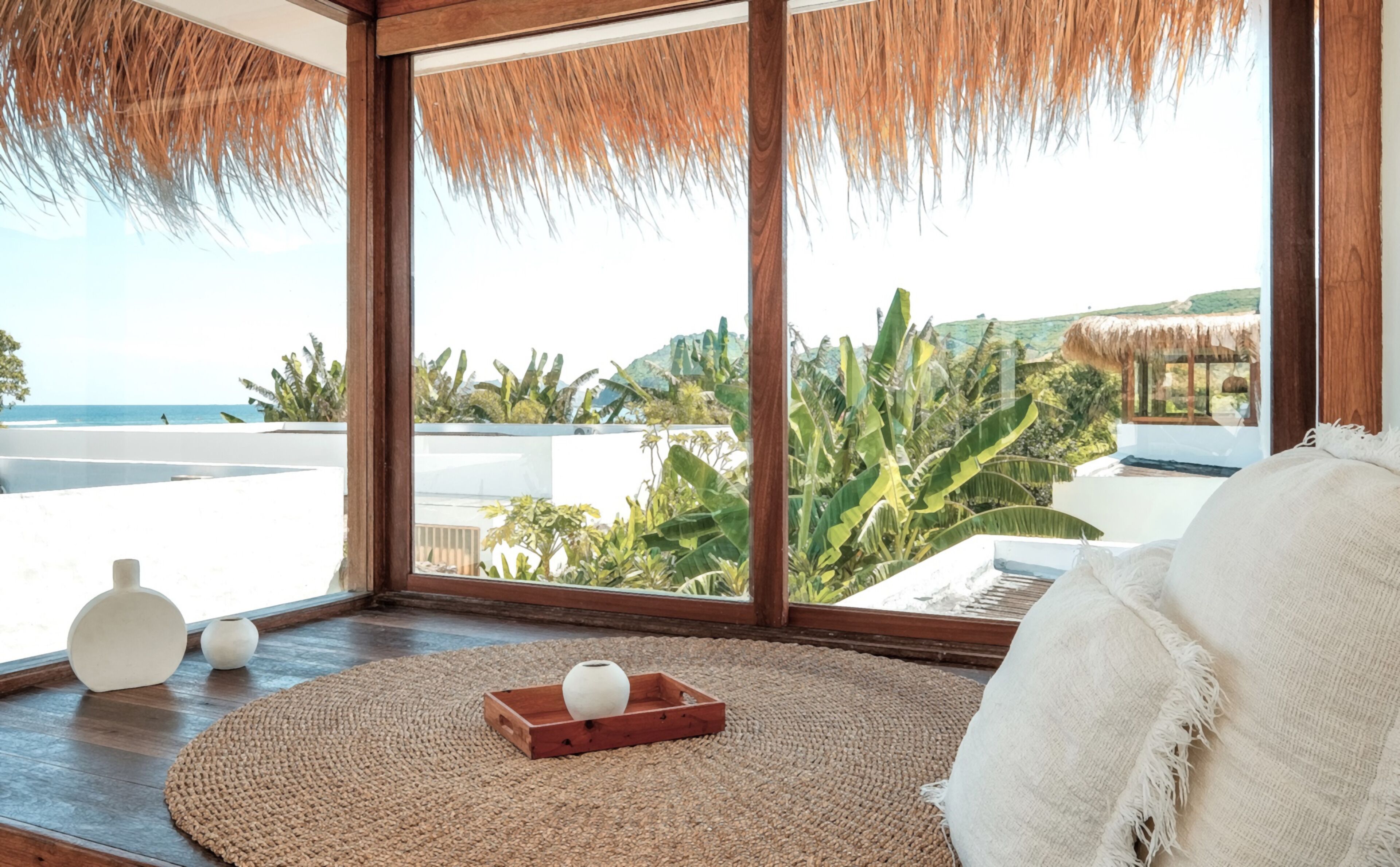 Una habitación con un ventanal grande, un tapete tejido, una bandeja de madera con un florero y una almohada blanca.