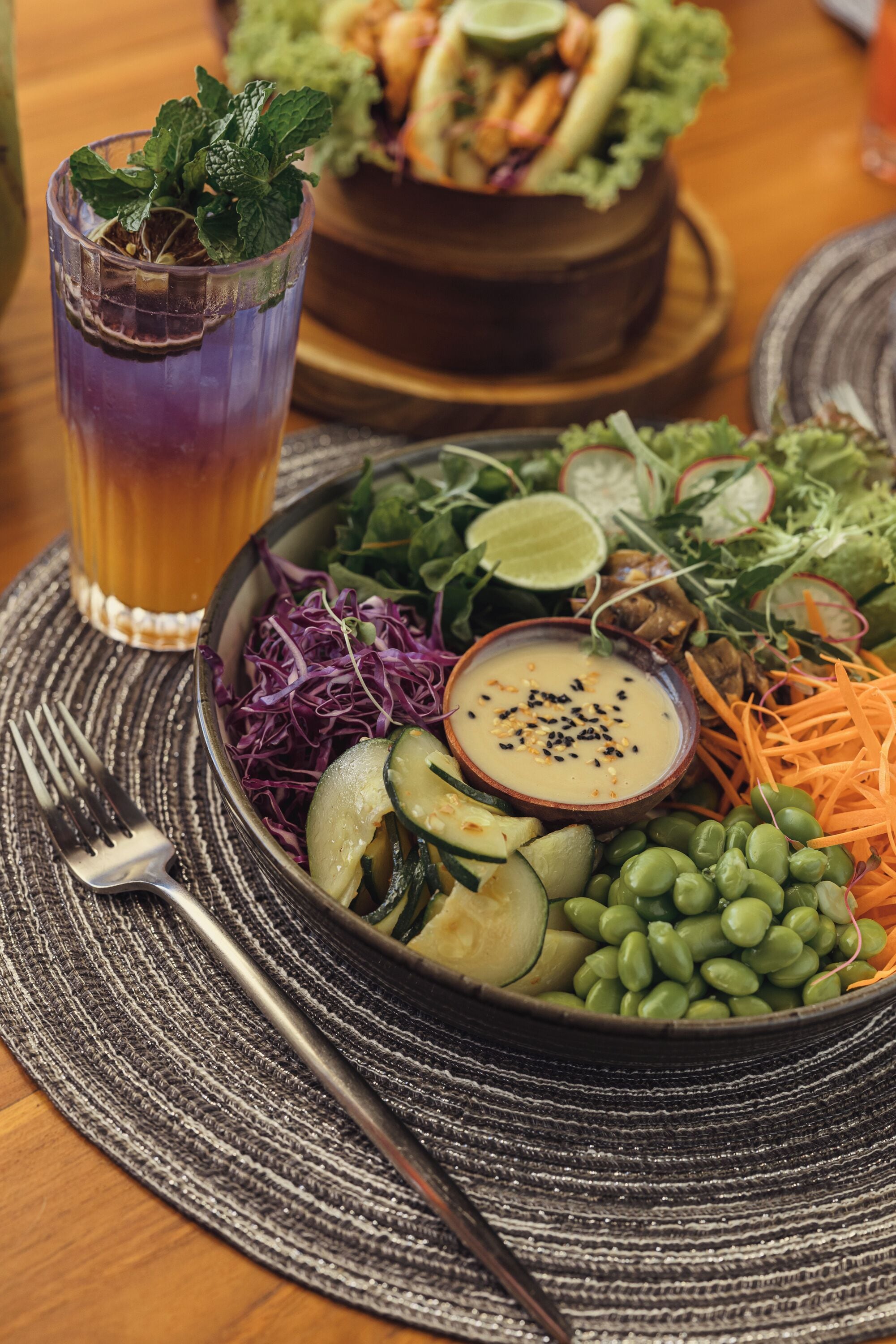 Un plato con diferentes verduras, un tazón de salsa y un vaso de bebida sobre una mesa.