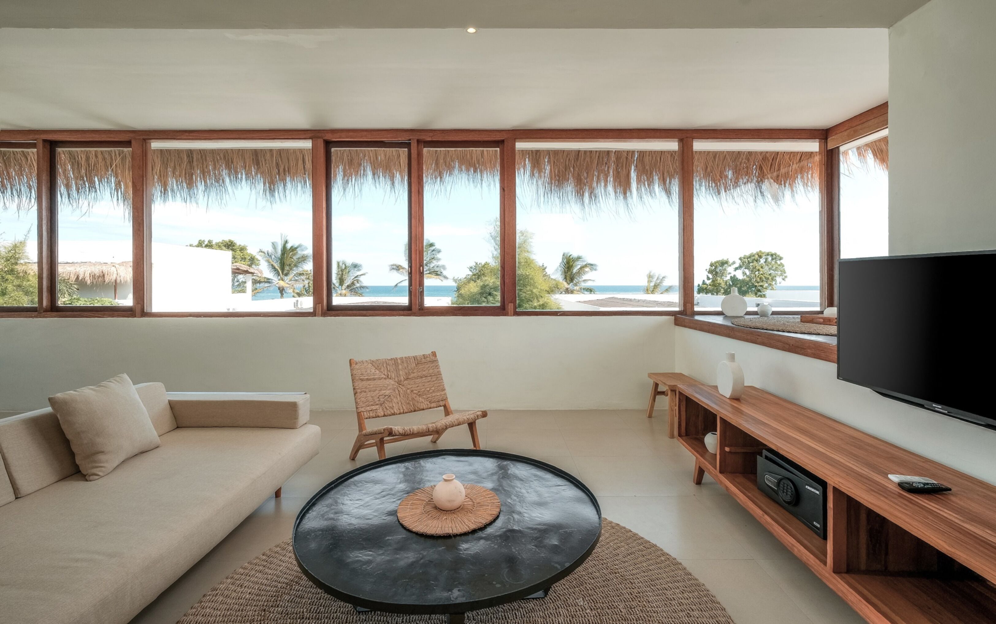 A modern living room with a sofa, a round coffee table, a TV, and a large window overlooking a beach.