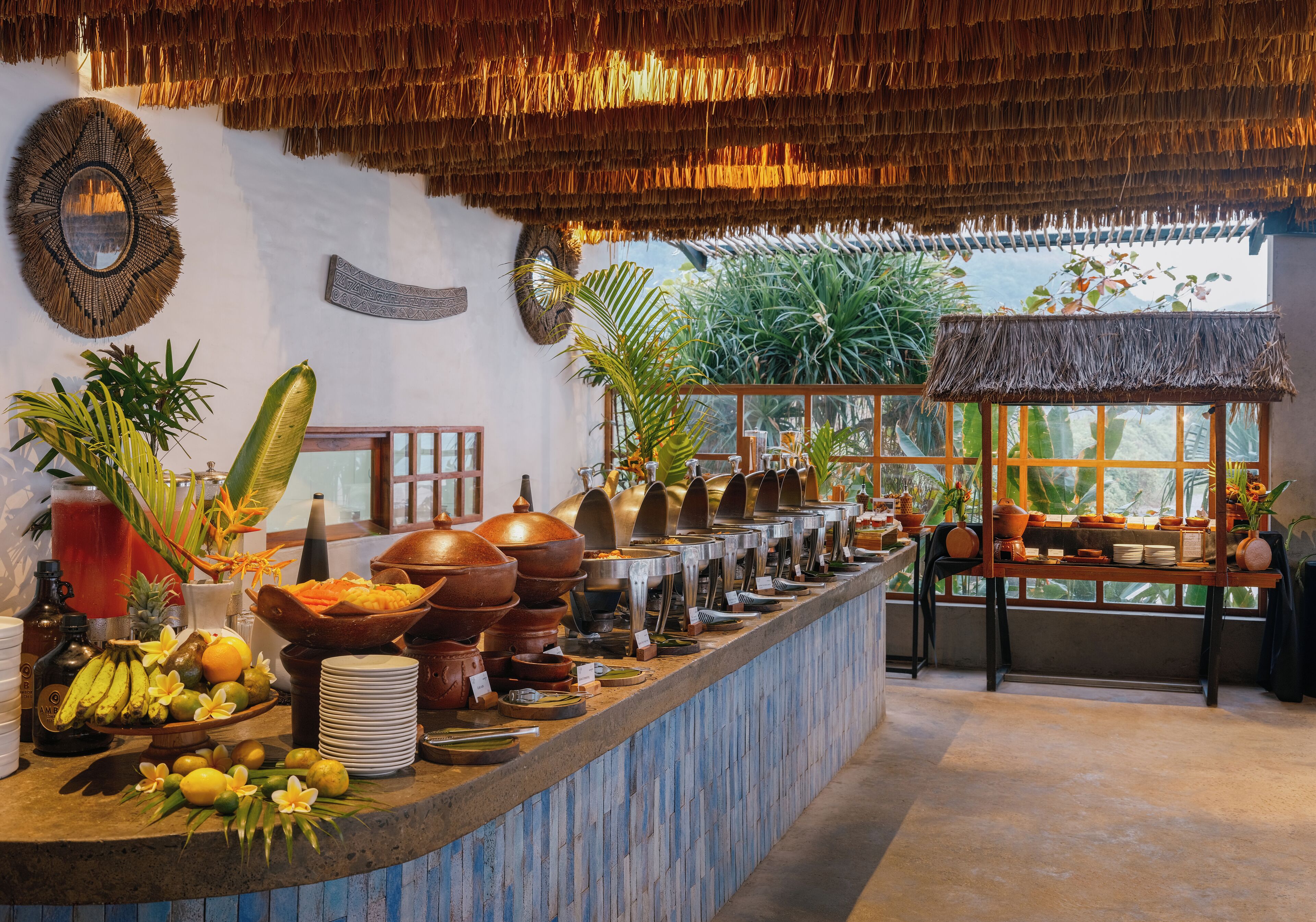 A buffet area with a variety of dishes and drinks, a thatched roof, and tropical plants.