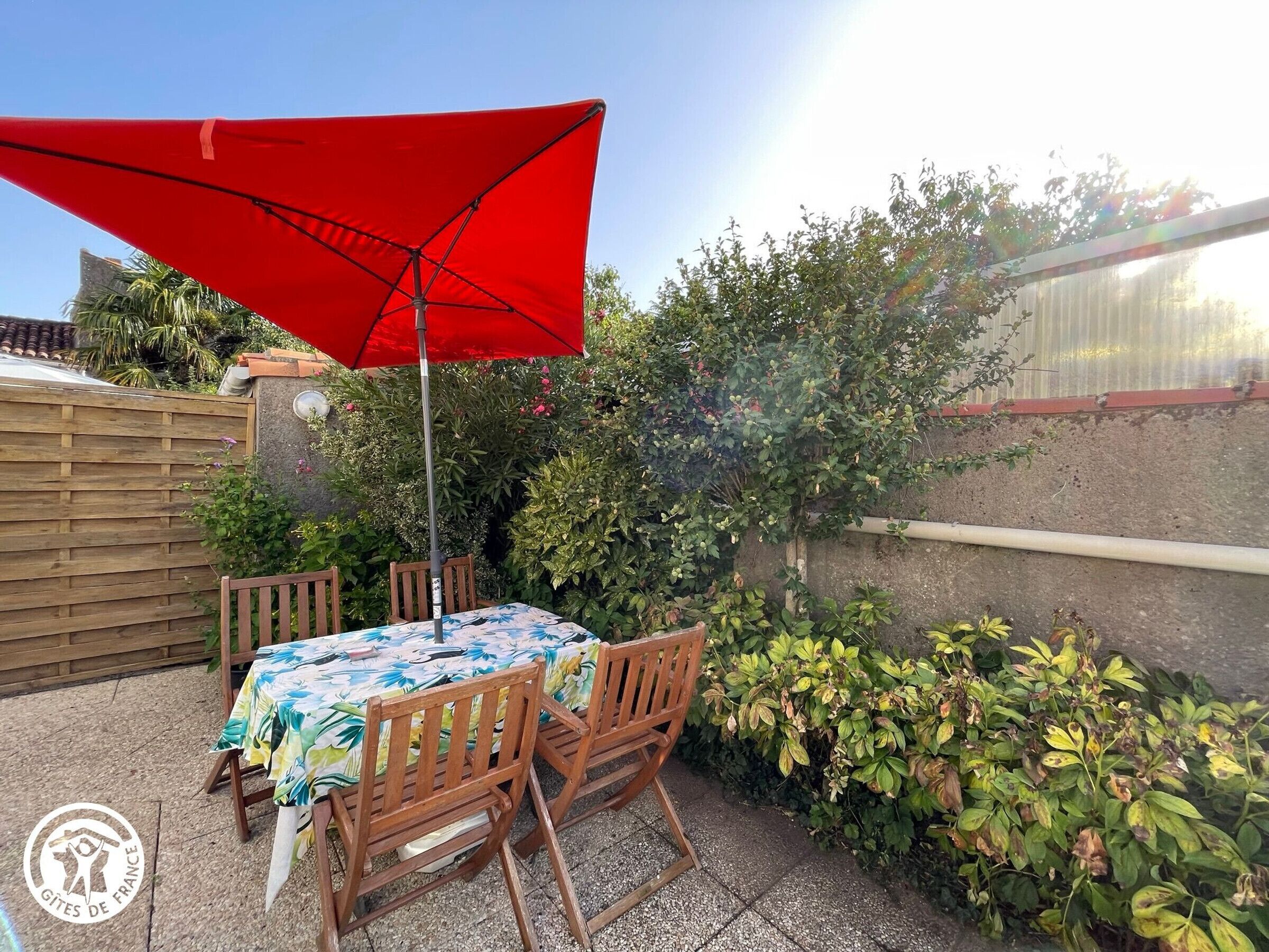 Een terras met een tafel en stoelen onder een rode parasol, omringd door groen en een houten hek.