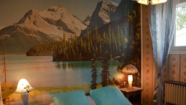 Ein Schlafzimmer mit Wandgemälde von einer Berglandschaft, einem Bett mit zwei Kissen, einem Nachttisch mit Lampe und einem Heizkörper.