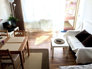 A living room with a wooden floor, a white sofa, a coffee table, a TV on a stand, and a large window with blinds. - Apartment Westside (Regensburg)