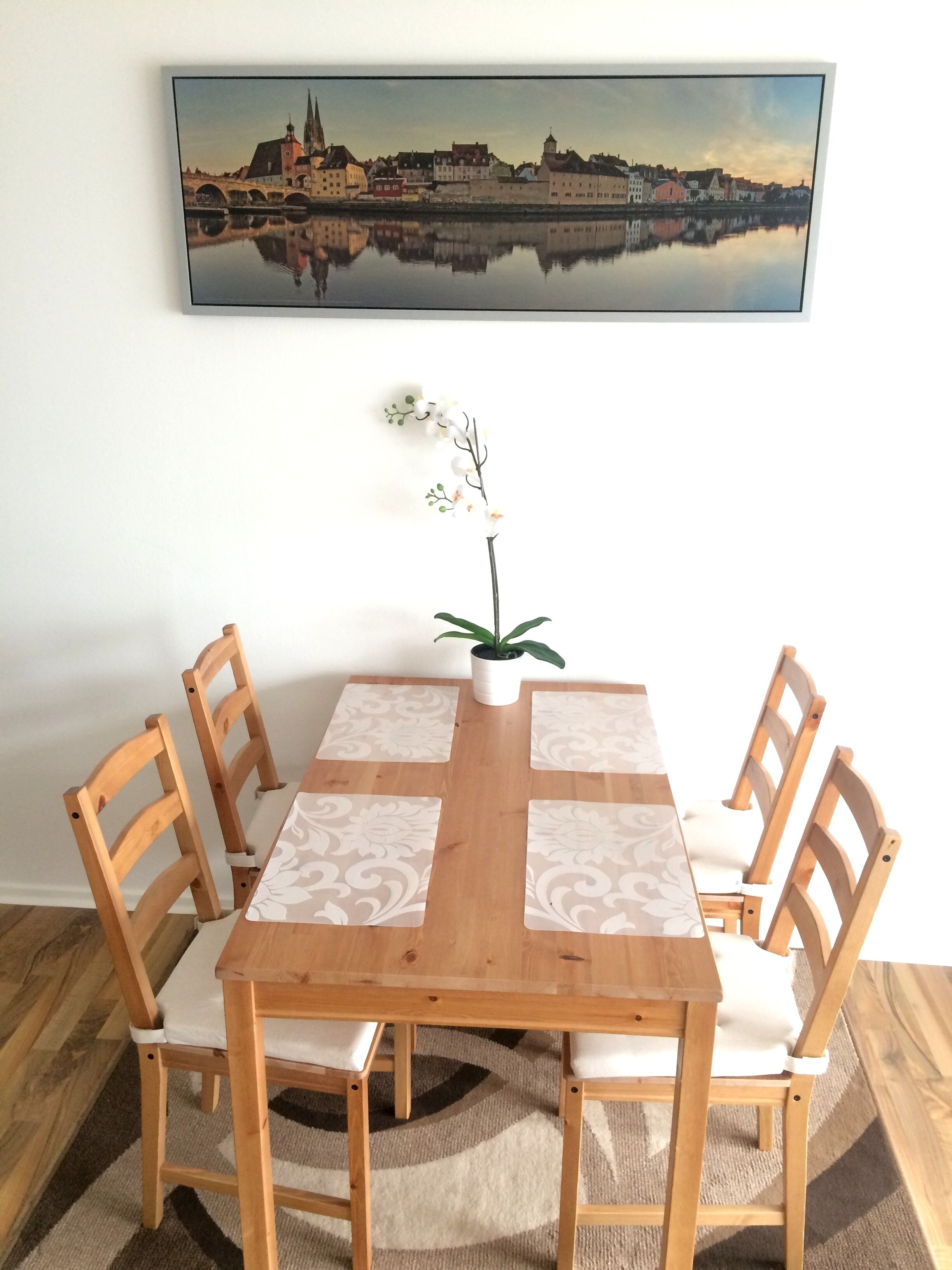 Una mesa de comedor con cuatro sillas, un florero con flores y un cuadro de un paisaje urbano en la pared.