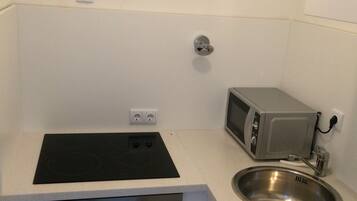 Una kitchenette compacta con cocina por inducción negra, horno integrado, microondas pequeño y pileta de acero inoxidable.