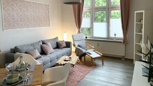 A living room with a grey sofa, a white armchair, a wooden coffee table, and a large window with curtains. - Premium city apartment * underground parking, Netflix, Prime * (Cottbus)
