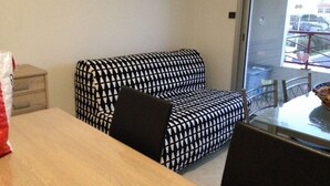 A room with a patterned sofa, a wooden table, and a window with a view of a building and parked cars. - Studio 100m from the Sea in Saint-Hilaire-de-Riez, with Balcony and Public Parking (Saint-Hilaire-de-Riez)