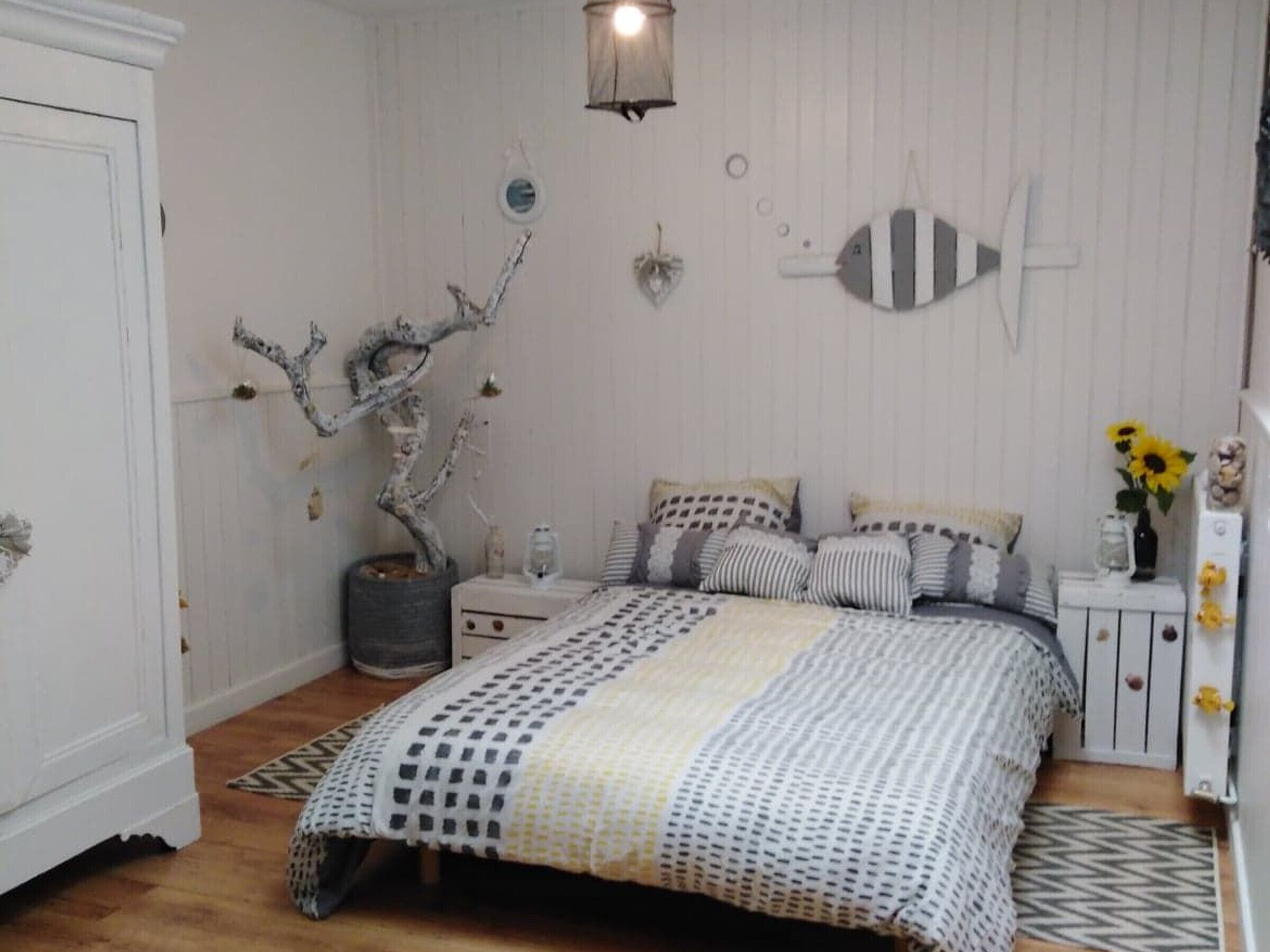 Ett sovrum med en säng, sängbord, en lampa, ett dekorativt träd och en spegel som hänger på väggen.