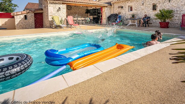 A swimming pool with inflatable toys, a stone building with a tiled roof, and a clear blue sky.