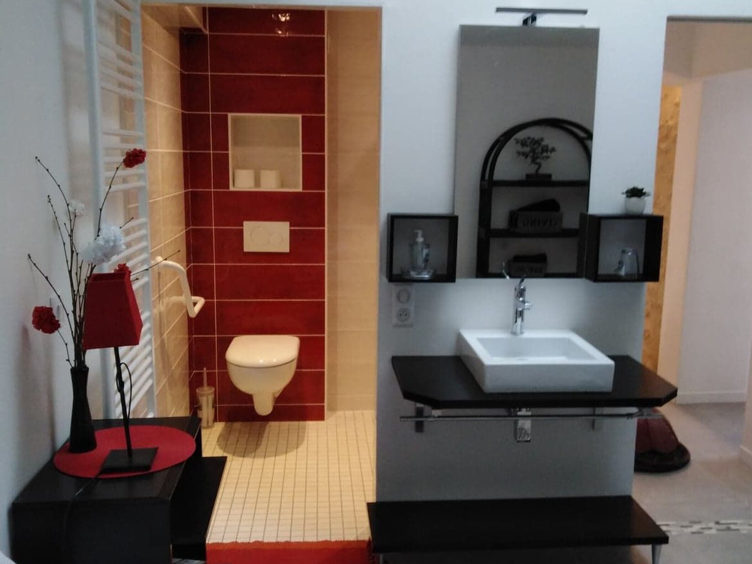 A modern bathroom with a red tiled wall, a white toilet, a black sink, and a mirror above it. There are decorative vases on the floor and a small shelf with toiletries.