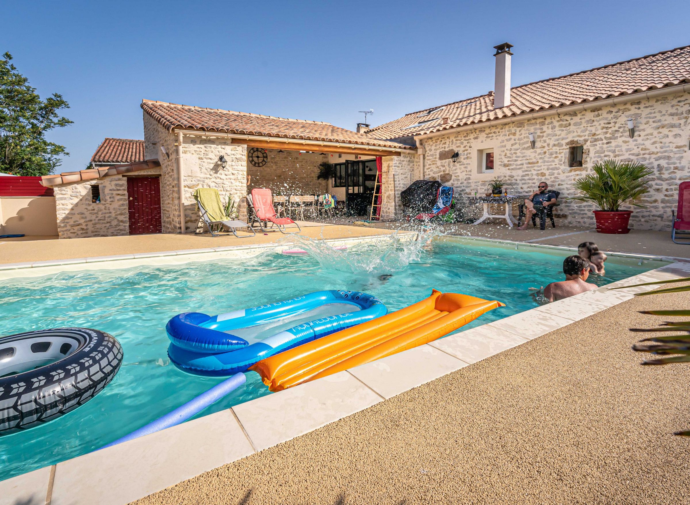 Una piscina con juguetes inflables, un edificio de piedra con techo de tejas y un cielo azul despejado.