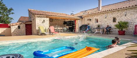Una piscina con juguetes inflables, un edificio de piedra con techo de tejas y un cielo azul despejado.
