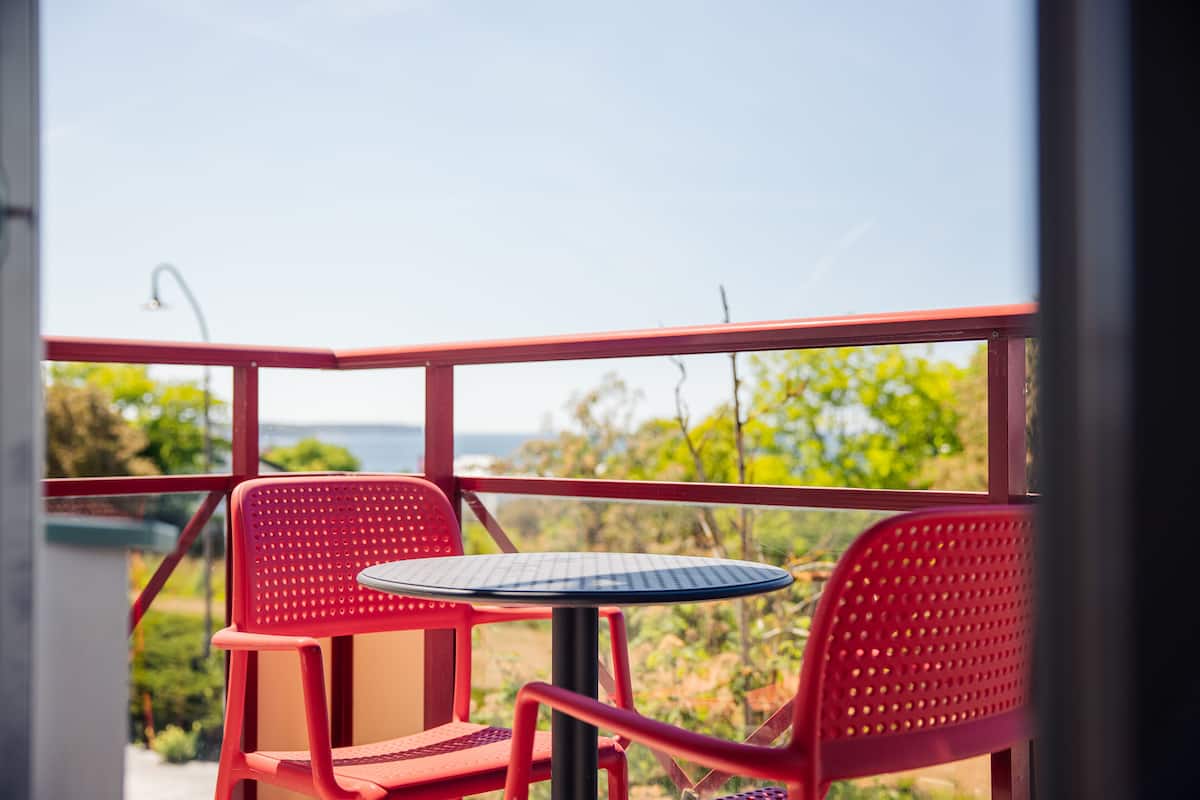 A red metal balcony with a round table and two chairs, overlooking a garden and a body of water.