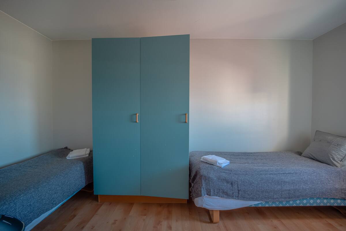 A room with a teal wardrobe, two single beds with grey bedding, and wooden flooring.