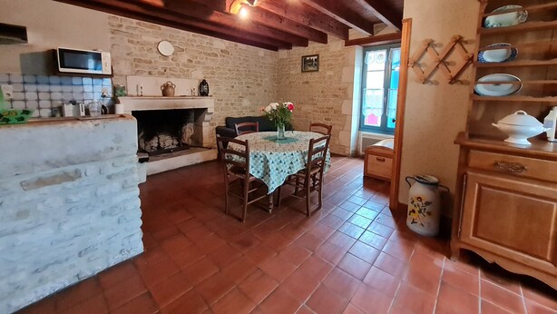 A rustic kitchen with a stone wall, wooden beams, a tiled floor, a dining table with chairs, and a wooden cabinet with dishes.