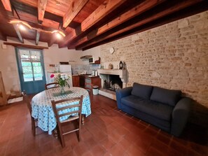A cozy living room with a stone wall, a fireplace, a blue sofa, and a dining area with a table and chairs. - Charming 1900s House in Marais Poitevin with Garden, Terrace, and Fishing Access (Benet)