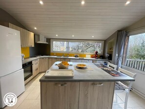 A modern kitchen with a central island, wooden cabinets, and a large window providing a view of the outside. - Cozy Riverside Cottage with Canoes, Terrace & Garden in Charming Damvix (Damvix)