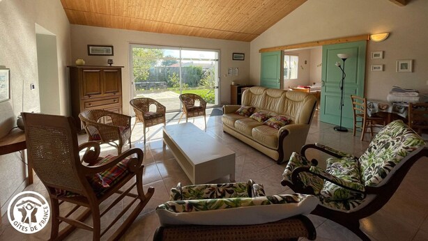Un salon spacieux avec un haut plafond en bois, un canapé, deux fauteuils, une table basse et une vue sur un jardin, accessible par une porte-fenêtre coulissante.