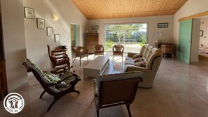 Un vaste salon doté d’un haut plafond en bois, d’une grande porte-fenêtre coulissante donnant sur une terrasse, et d’un coin salon confortable avec un canapé et des fauteuils.