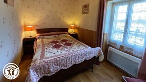 A bedroom with a wooden ceiling, a bed with a floral quilt, two bedside lamps, a window with curtains, and patterned wallpaper. - Rustic cottage with games room and animals in the countryside (Saint-Julien-des-Landes)