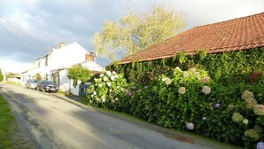 A quiet street with a house featuring a red-tiled roof and a lush green hedge. - Rustic cottage with games room and animals in the countryside (Saint-Julien-des-Landes)