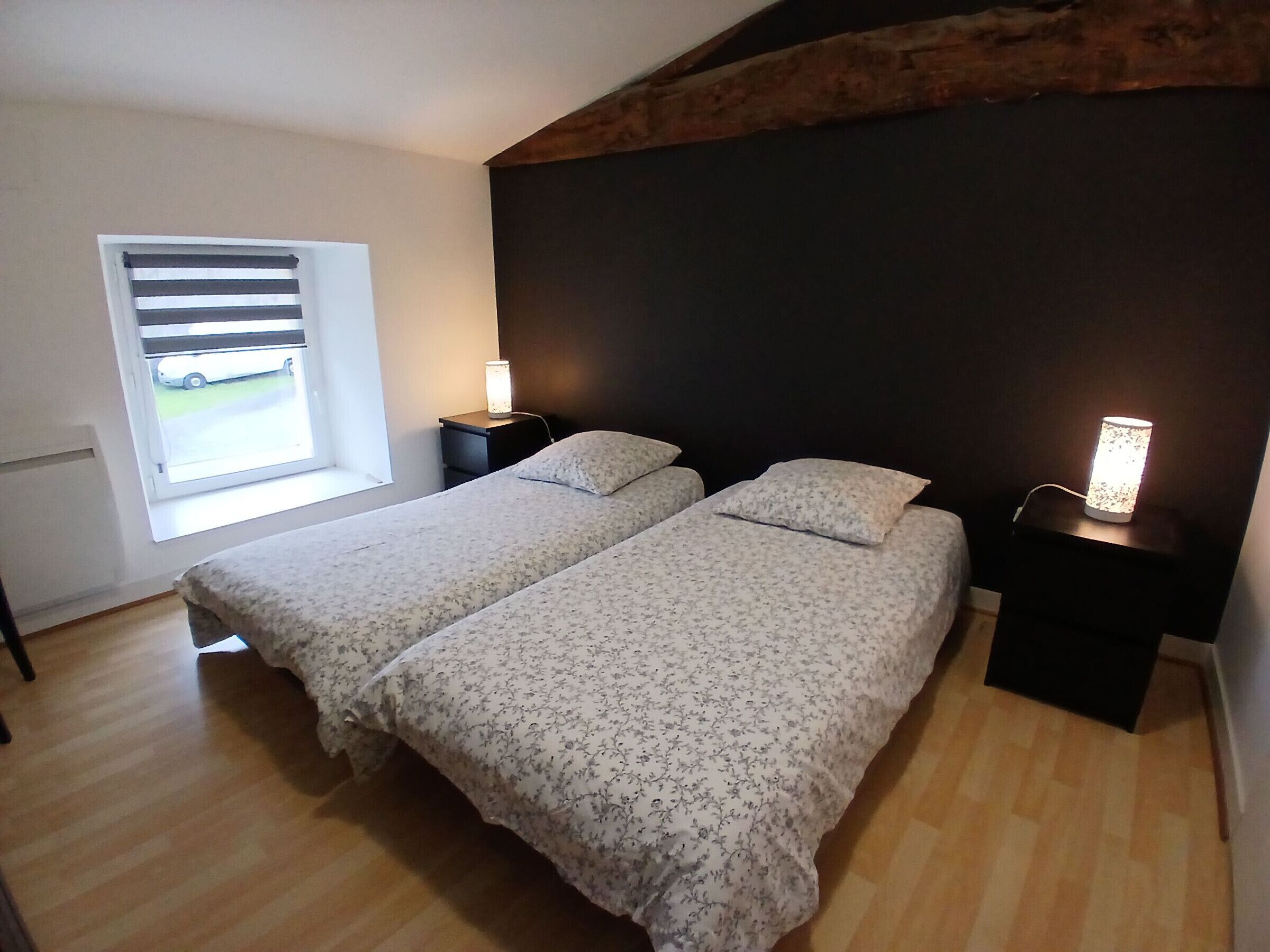 A bedroom with two beds, a window with blinds, a nightstand with a lamp, and a wooden ceiling beam.