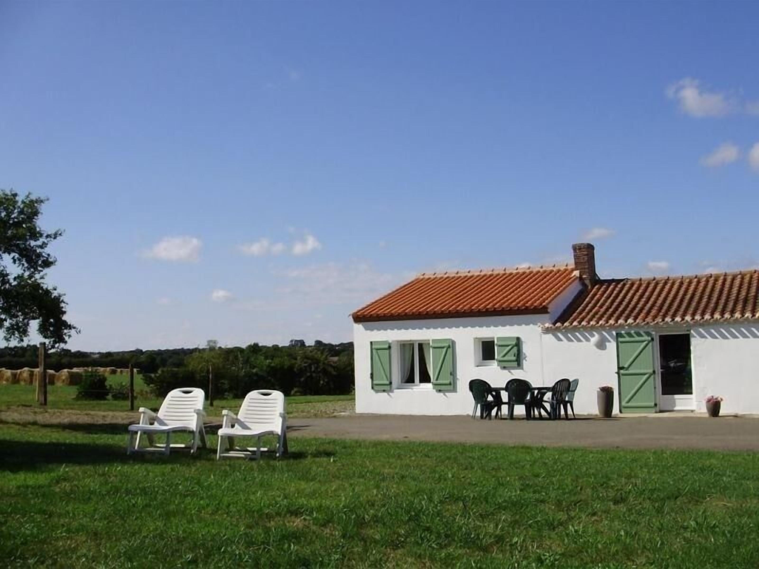 A small white building with a red-tiled roof, green shutters, and a patio with outdoor furniture.