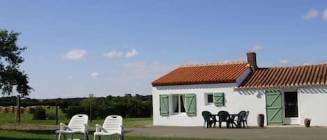 A small white building with a red-tiled roof, green shutters, and a patio with outdoor furniture.