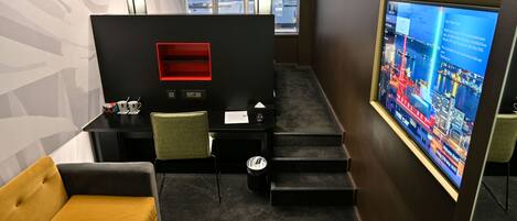 A modern hotel room with a black partition, a yellow sofa, a small table with a trash bin, and a large TV screen displaying a cityscape.