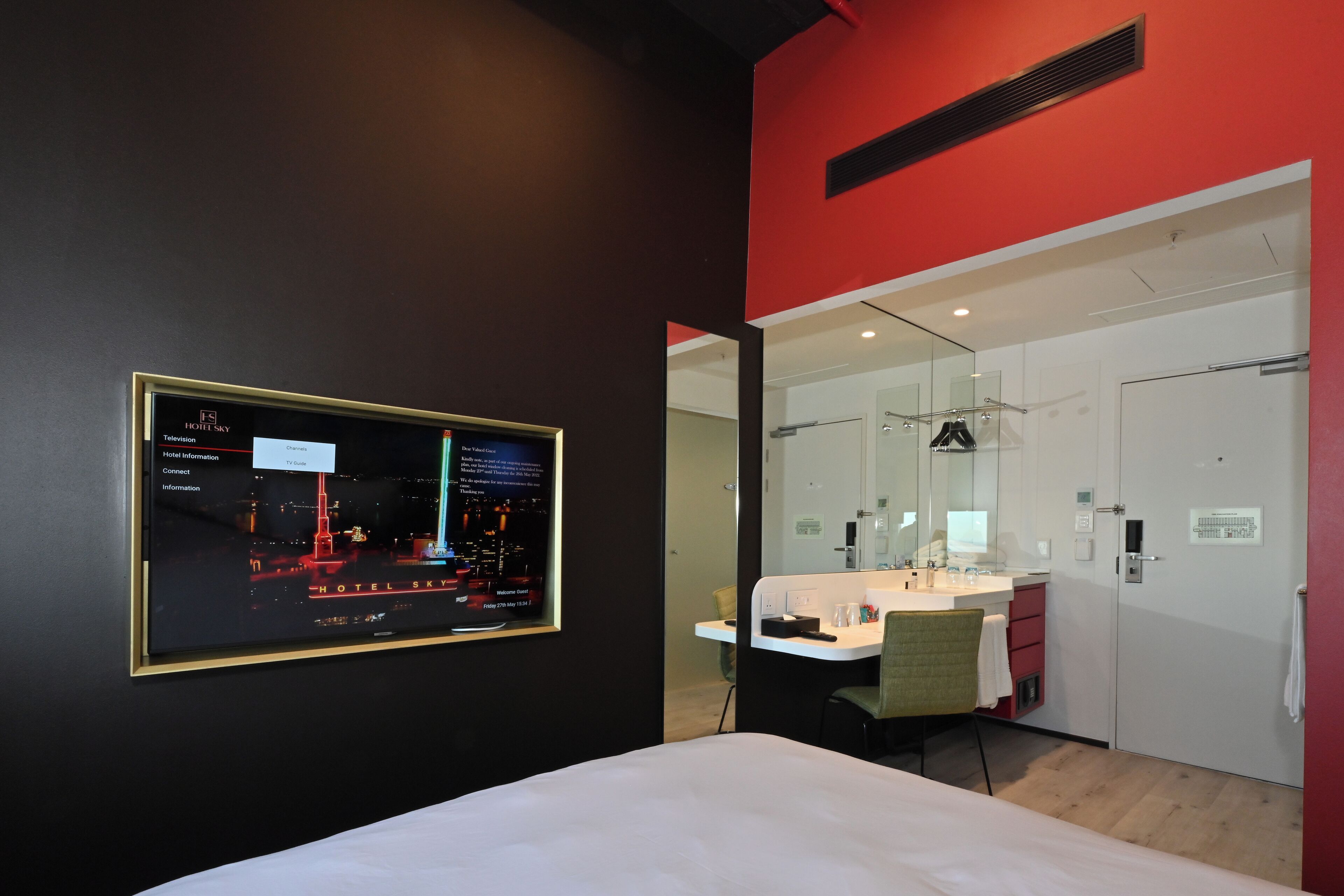 A hotel room with a bed, a desk, a chair, a TV displaying hotel information, and a bathroom with a mirror and sink.