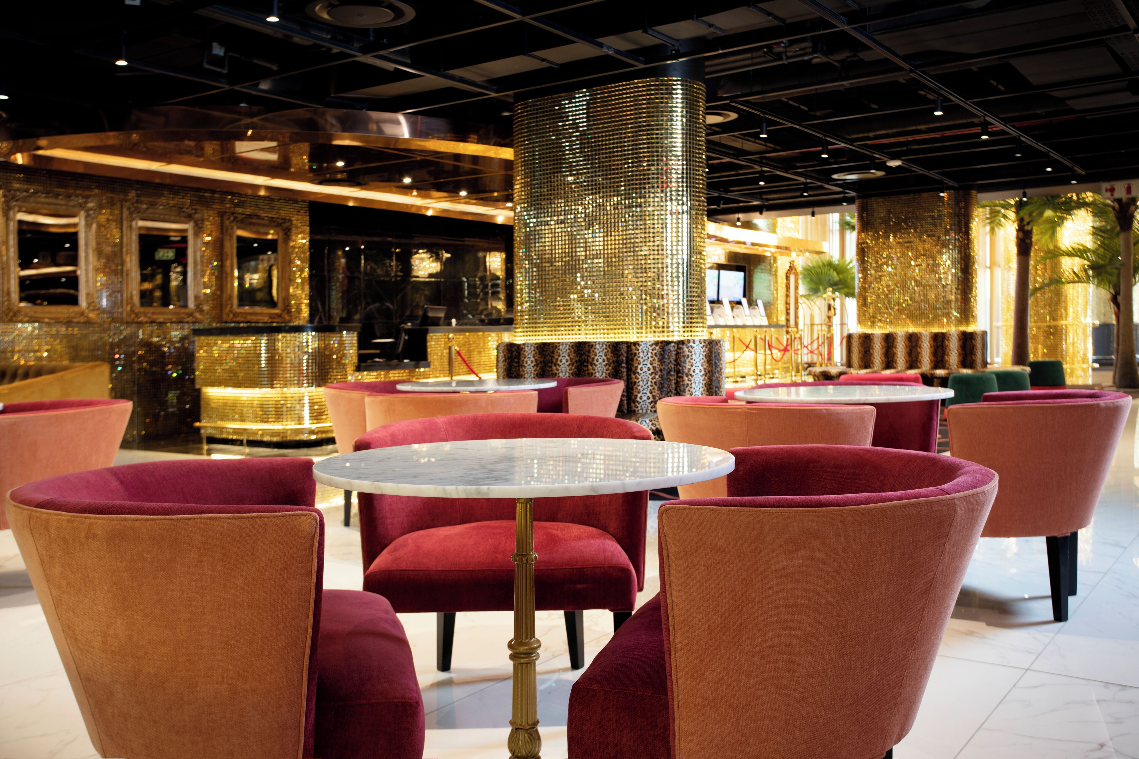 Ein modernes Interieur mit runden Tischen und bequemen roten Sesseln, einer dekorativen Goldkolonne sowie einer Bar mit Spiegel und Pflanzen.