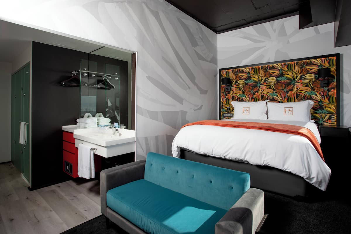 A modern hotel room with a bed, a sofa, a bathroom with a mirror and towel rack, and a wall with a tropical leaf pattern.