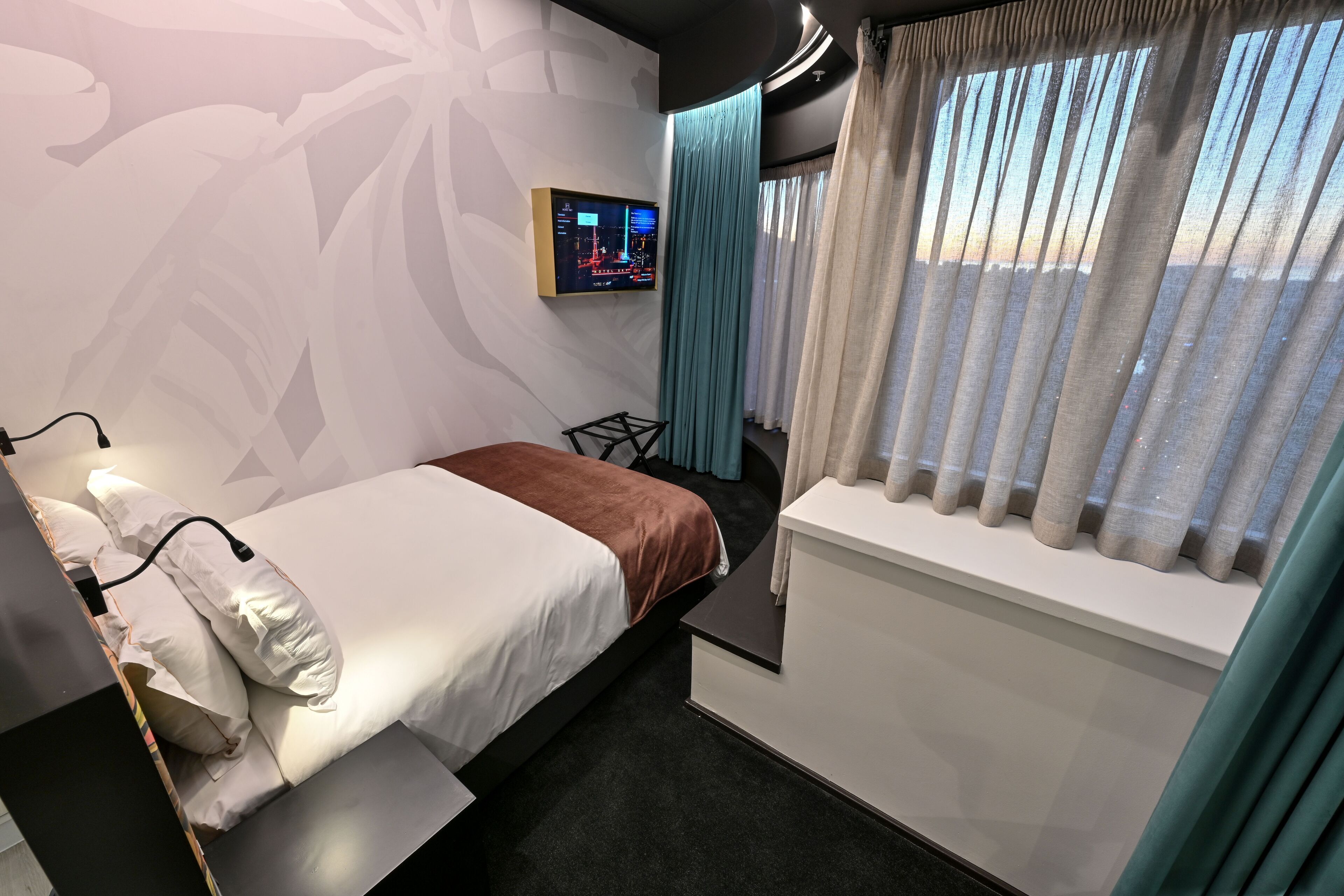 A modern hotel room with a bed, a small desk, a TV on the wall, and a window with curtains.