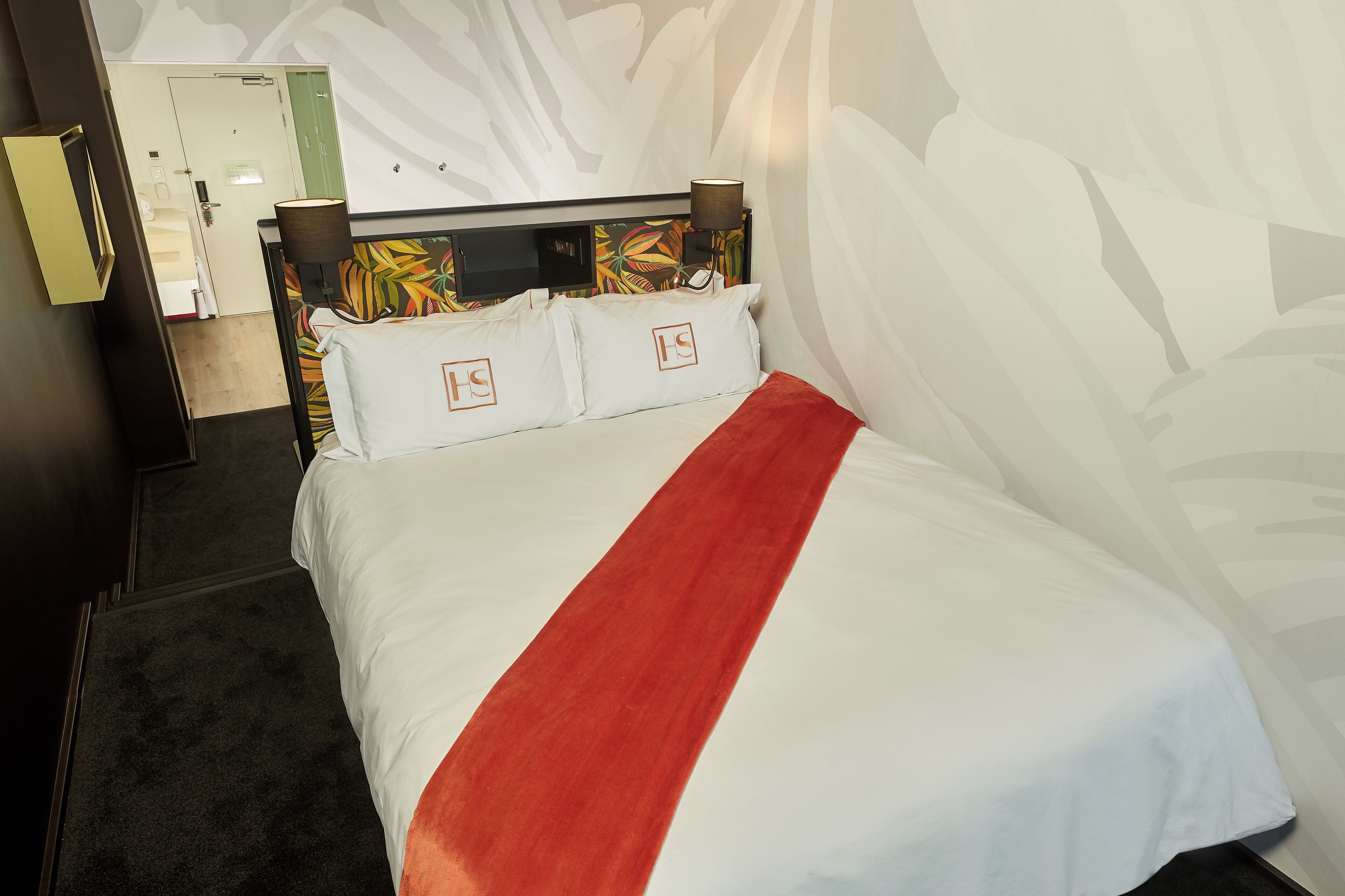Una habitación de hotel con cama, cabecera con estampado floral, manta roja y pared con diseño de hojas.