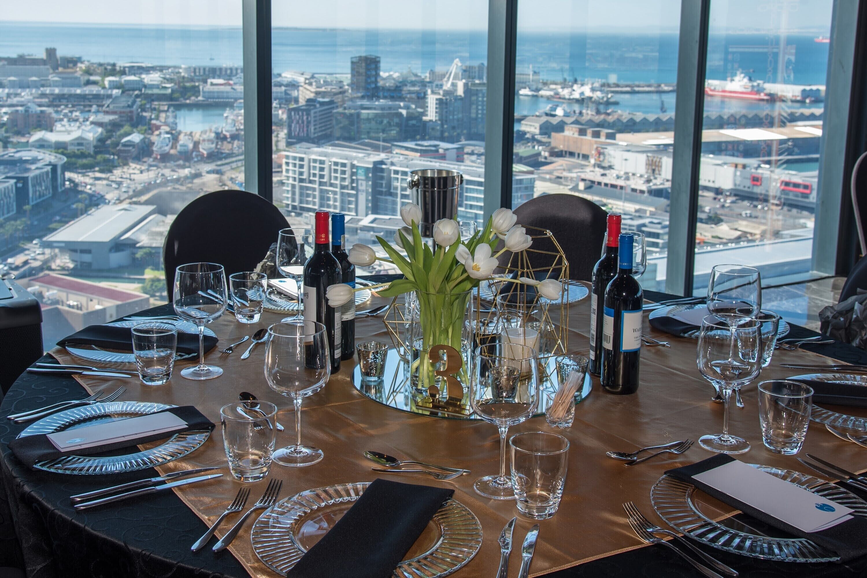 Una mesa lista para comer con vista a la ciudad, un florero con flores blancas y botellas de vino.