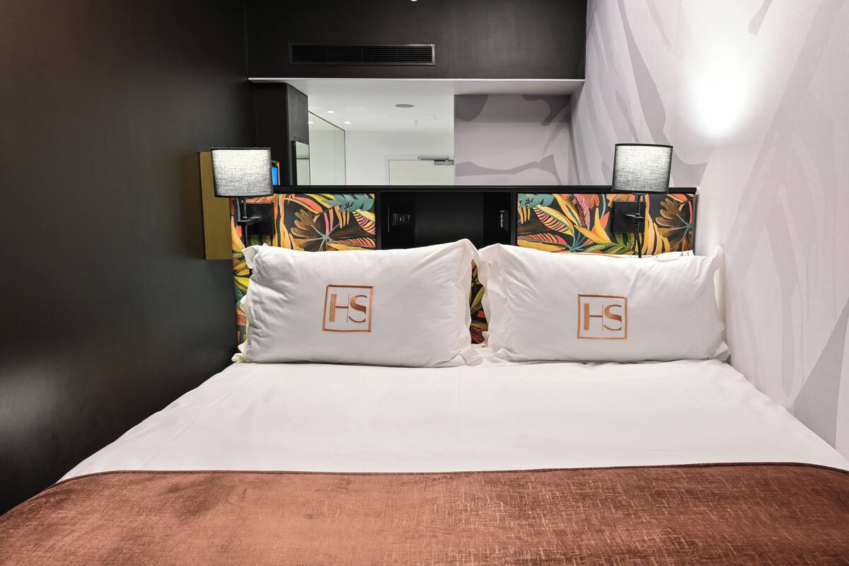 A hotel room with a large bed, two white pillows with a logo, a nightstand with a lamp, and a mirror above the bed.