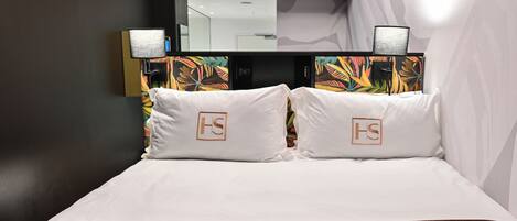 A hotel room with a large bed, two white pillows with a logo, a nightstand with a lamp, and a mirror above the bed.