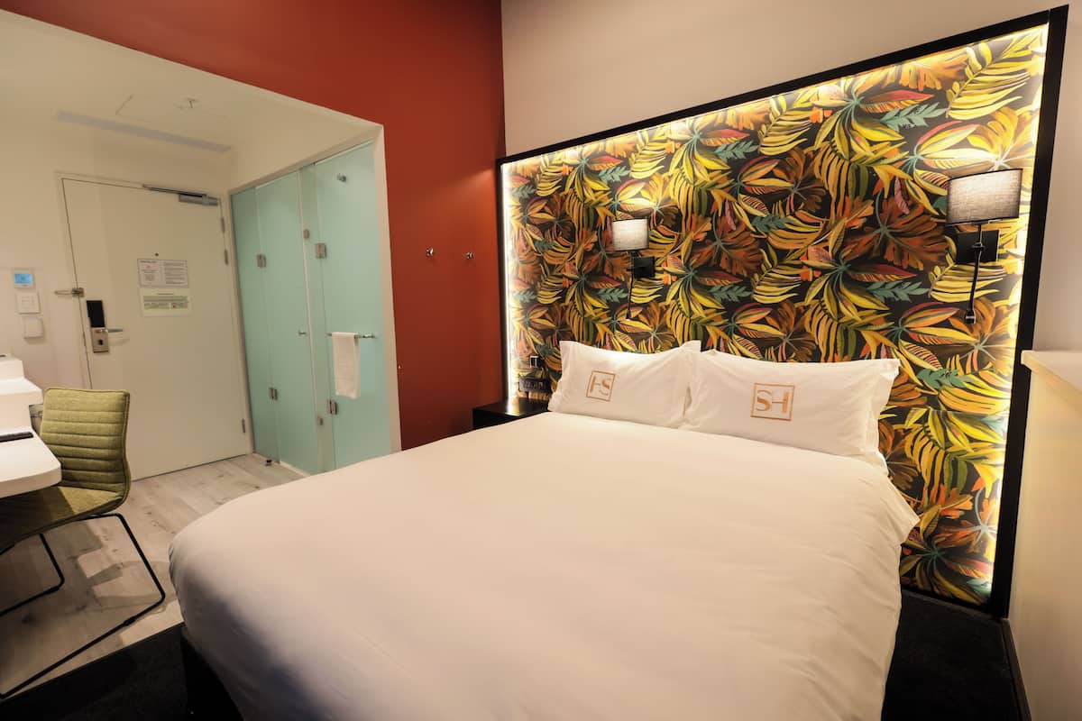 A hotel room with a bed, a desk, and a chair. There is a door and a wall with a leaf pattern.