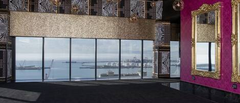 Una habitación amplia con un gran ventanal que ofrece vistas a un puerto y barcos, una pared con diseño y un espejo con marco ornamentado.
