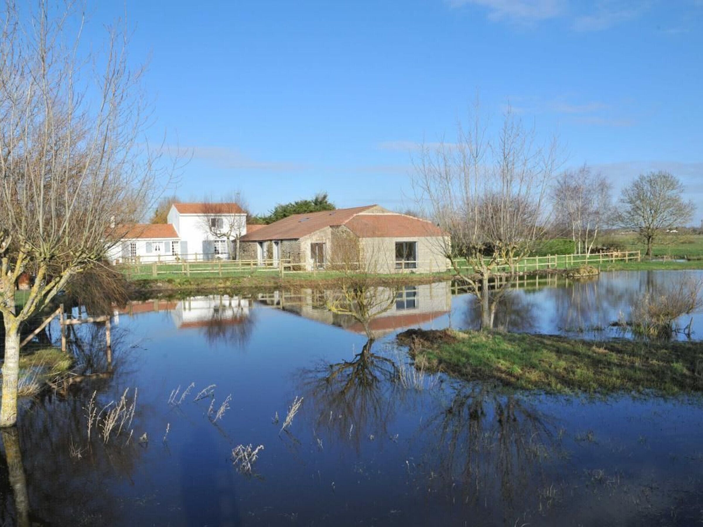 Une maison avec un toit rouge et des murs blancs, un étang au premier plan et un ciel bleu dégagé.