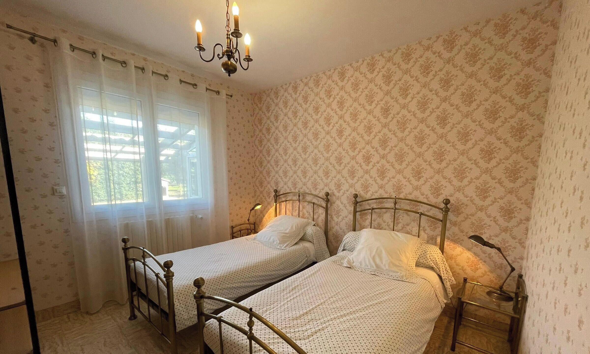 Une chambre avec deux lits simples, un lustre, une fenêtre avec des rideaux et un papier peint à motifs.