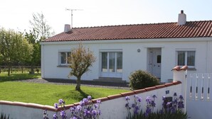 A white building with a red-tiled roof, a small tree, and purple flowers in the foreground.
