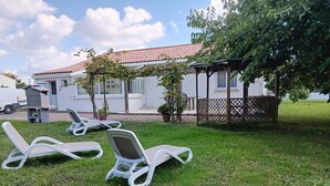 A grassy area with three white lounge chairs, a white building with a red-tiled roof, and a wooden pergola.